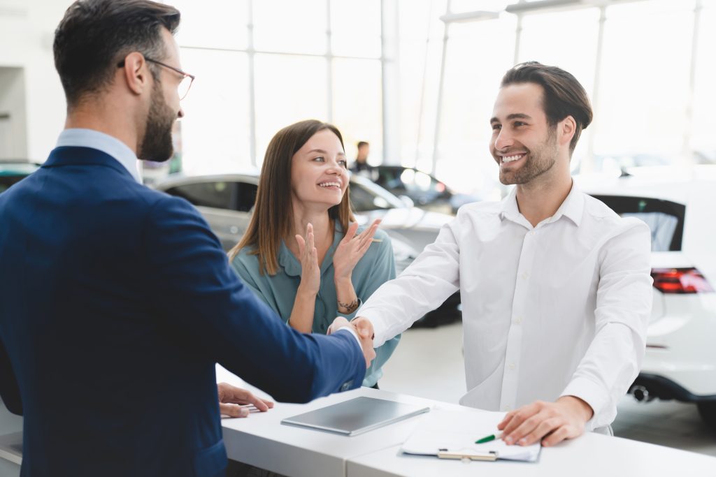 In a GMC showroom sales professional shakes hands with a smiling new couple who have just finished signing on the dotted line of their financial agreement while the woman claps her hands