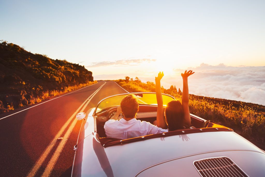 We see a couple from behind driving a sports car with the top down beside a lake at sunrise while the woman raises her hands into the air with worry-free joy